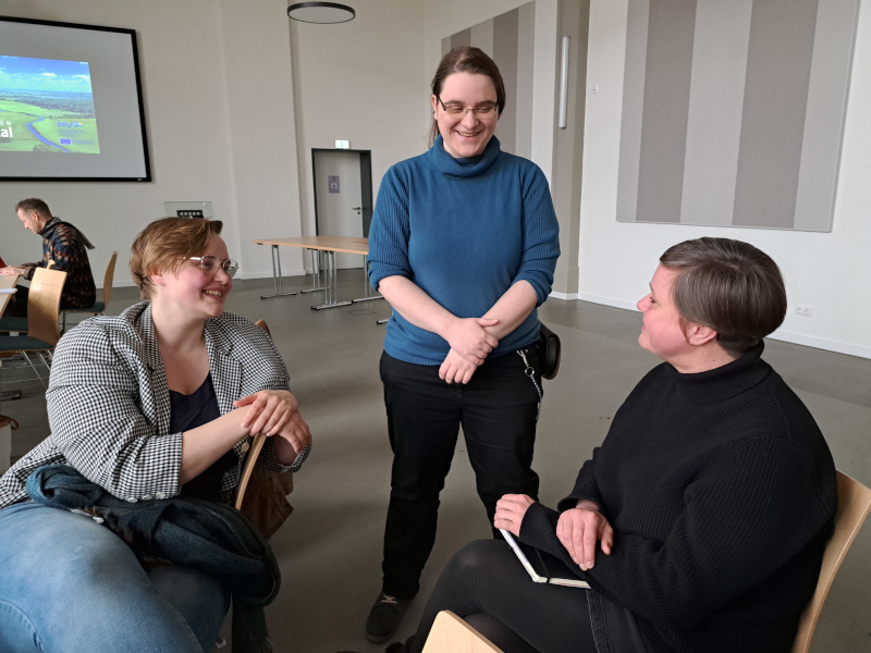 Gesprächsrunde Anna Goedicke, Anne Zandt und Anja Seegert (im Hintergrund Thomas Radke erkennbar). Anne steht zwischen den beiden sitzenden anderen, sie lachen gemeinsam