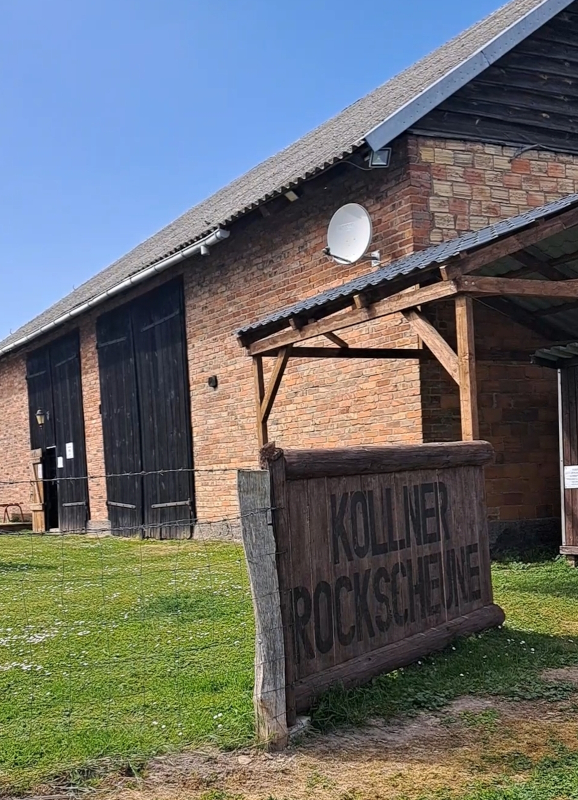 Foto der Rockscheune. Vorne ein großes Holzschild mit dem Schriftzug "Köllner Rockscheune" dahinter die Scheune selbst mit offenem Tor