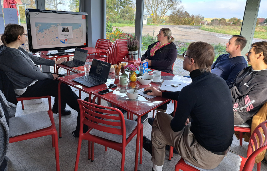 Foto vom Karten- und Kalenderworkshop, mit fünf Personen, rotem Tisch und roten Stühlen, Laptops und einem großen Fernseherbildschirm auf dem gerade eine Karte präsentiert wird.