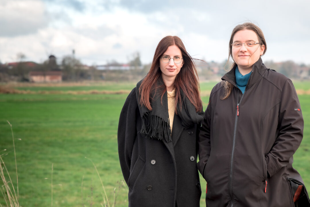 Pia Schulze und Anne Zandt von der Kulturregion Tollensetal stehen auf einem grünen Feld vor der verschwommenen Kulisse von Alt Tellin und lächeln in die Kamera.